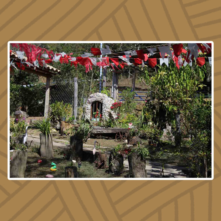 Para todos verem: Um quintal ensolarado, com um caminho decorado por troncos de árvores com plantas ornamentais e bibelôs de jardim em suas margens. Bandeirinhas de festas juninas, vermelhas e brancas, sombream todo o caminho. No fim do percurso, ao centro da imagem, uma gruta oval, de acabamento em pedras de cascalho em tons claros, contrastam com o interior escuro onde se abriga uma imagem do santo de devoção. Hastes finas de ferro compõem o portão da gruta.