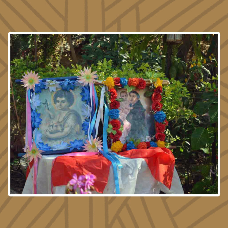 Para todos verem: Em uma mesa, duas bandeiras ornadas com moldura de flores e fitilhos nas cores azul-claro, branco e rosa. A bandeira da esquerda traz consigo a imagem de São João Batista, sendo ele um menino que carrega um cordeiro nos braços. A moldura é composta por flores azuis e brancas. A bandeira da direita traz a imagem de Santo Antônio, com seu traje de túnica franciscana marrom. Ele é um jovem homem calvo, carregando o menino jesus no colo. A moldura é composta por flores de rosas amarelas, vermelhas, azuis e laranjas.