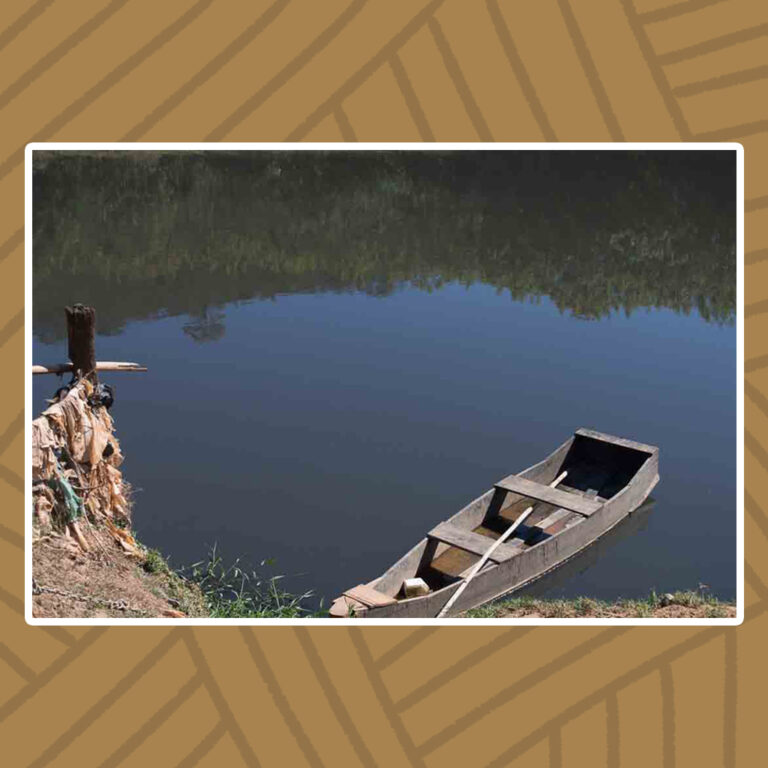 Para todos verem: O Rio das Velhas, de águas escuras, revelam através do reflexo da superfície , a margem oposta de vegetação curta de pequenos arbustos e céu aberto. Na parte inferior da imagem se encontra a margem com um barco de madeira. Seu interior possui uma haste de remo atravessada entre dois assentos de tábua de madeira. Há água no fundo da embarcação.