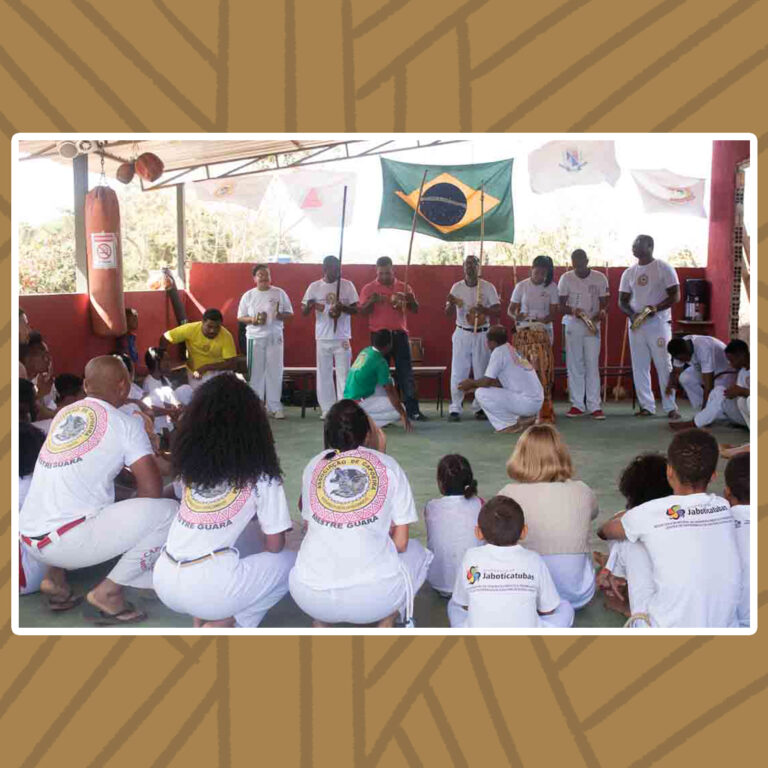 Para todos verem: Cena do jogo de Angola. Roda de capoeira formada por capoeiristas trajados com vestes brancas. Ao fundo, o grupo de percussão está de pé. Eles tocam xique-xique, pandeiros e berimbaus. Acima da percussão, estende-se uma bandeira do Brasil. Ao centro da roda, dois capoeiristas agachados se encaram um de frente para o outro.