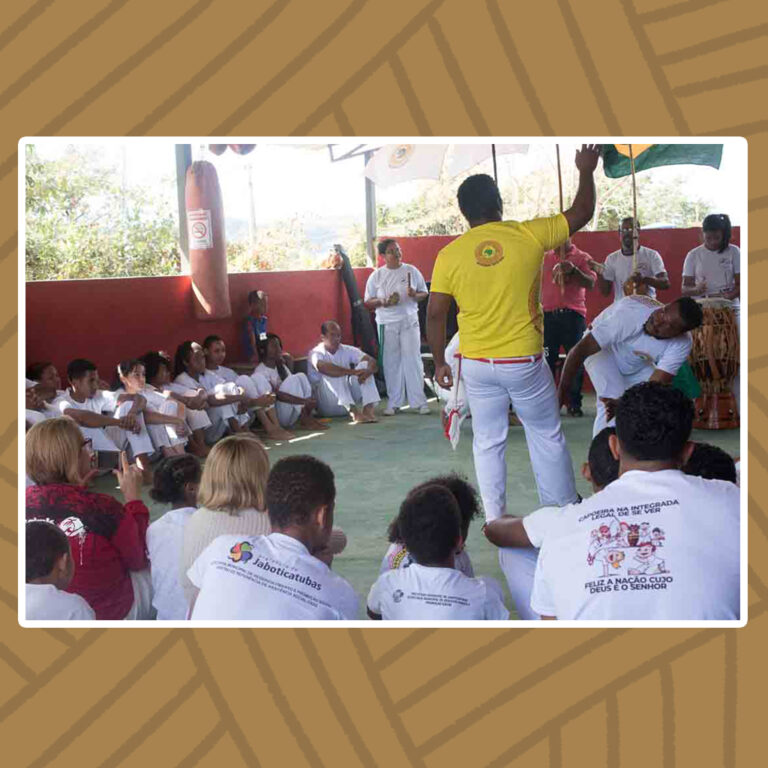 Para todos verem: Roda de capoeira. Jovens com trajes brancos de capoeira formam uma roda, alguns sentados no chão, outros agachados. O mestre da roda está de pé ao centro da roda. Ele se diferencia dos demais por estar usando uma camisa de malha amarela. Ele olha para o lutador que ginga à sua frente, além dos percussionistas da roda que estão de pé, com tambores e berimbau .