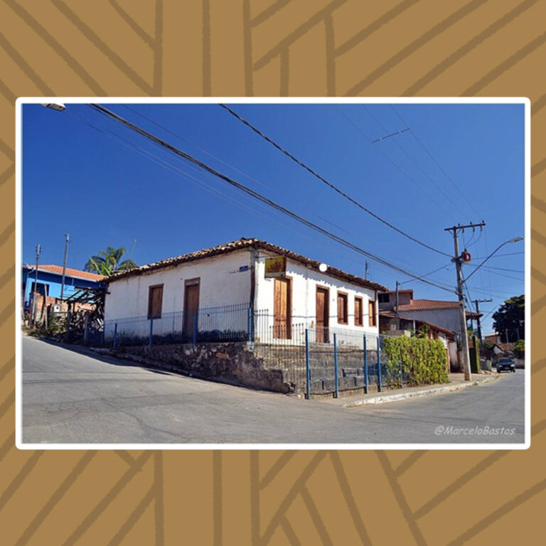 Para todos verem: Casarão colonial, de andar único, de telhado triangular simples com telhas coloniais, paredes brancas, portas e janelas de madeira rústica sem pintura. Uma escadaria de cascalho e concreto de quatro degraus leva à entrada principal. Nela, estão duas portas seguidas de três janelas à direita. Há uma entrada na lateral esquerda do casarão, composta por uma porta e uma janela. Uma cerca de grades azuis delimita o casarão, que está localizado entre o cruzamento de duas ruas.