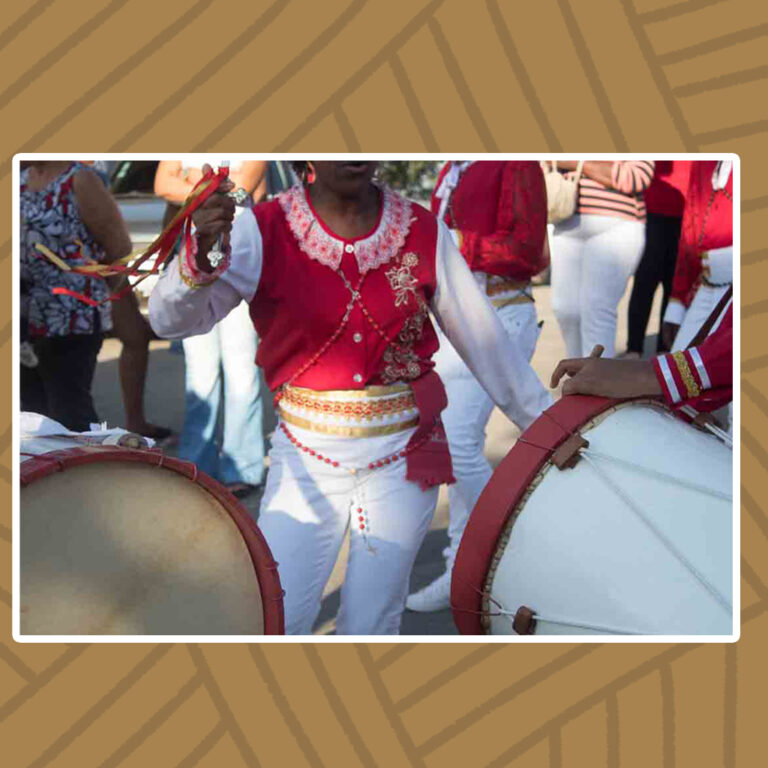 Para todos verem: Entre tambores e a multidão, uma mulher com trajes do Congado do Divino dança. Seu rosto é visto somente do queixo até a boca. Sua pele é negra, usa camisa vermelha de mangas longas e brancas com bordados. Usa calça branca, cinto dourado envolto na cintura e terços de contas rosas. Em uma das mãos segura fitas vermelhas e douradas.