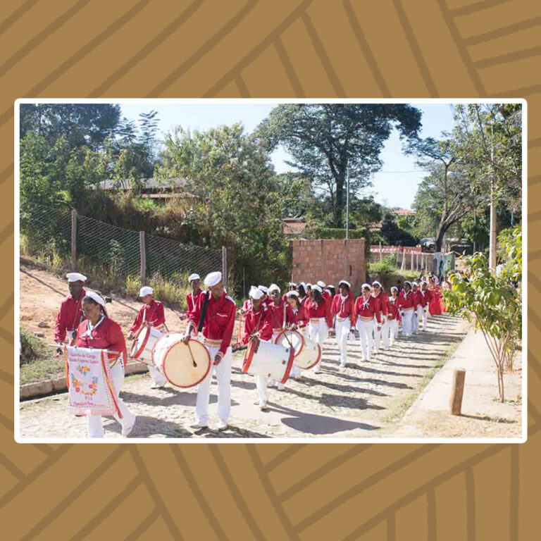 Para todos verem: Perfilados em pares, integrantes do Congado do Divino sobem uma rua íngreme. À frente da fila, uma mulher segura o estandarte do Congado do Divino. O congado é paramentado por vestes vermelhas e brancas: Chapéu de aba curta branco, Camisa vermelha de mangas longas e ombreiras brancas acompanhadas de laço branco no pescoço, cinta dourada e calça branca.