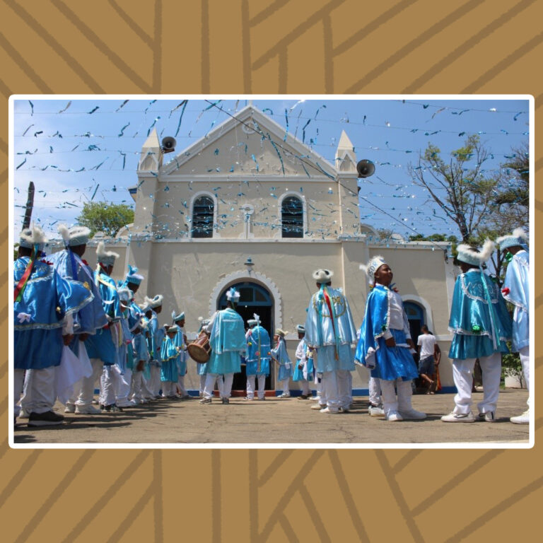 Para todos verem: Em um dia ensolarado, abaixo de um varal de fitilhos azul-claros e brancos, o Congado de Nossa Senhora do Rosário prepara a procissão de entrada. São duas filas de pares e tambores ao centro. Os integrantes usam manto azul-claro, chapéu camisa e calças brancos. A fachada da capela é triangular com dois obeliscos curtos nas extremidades. Na fachada de parede cor creme, tem janelas e portas azuis de arcos de dormentes brancos, além de suas marquises também em branco.