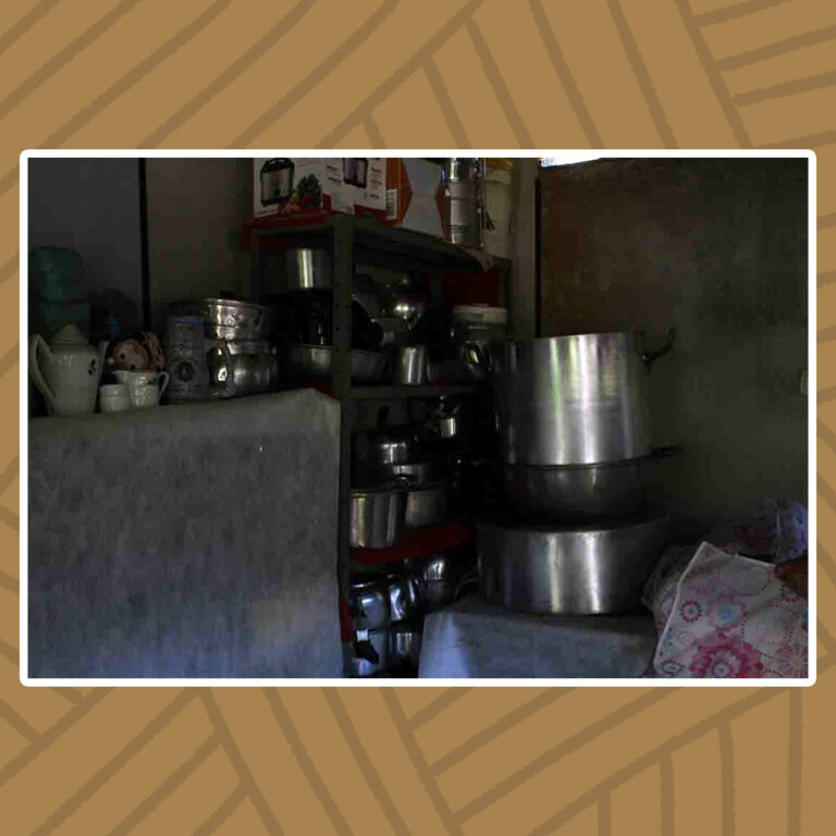 Para todos verem: Em uma cozinha coberta pela sombra do dia, brilha um conjunto de panelas, canecos e demais utensílios de metal. Todos empilhados em um armário de prateleiras abertas. À direita do armário, destaca-se uma pilha de panelas de tamanho industrial sobre uma mesa. À esquerda do armário, um conjunto de bule e xícaras de cerâmica em uma bancada.