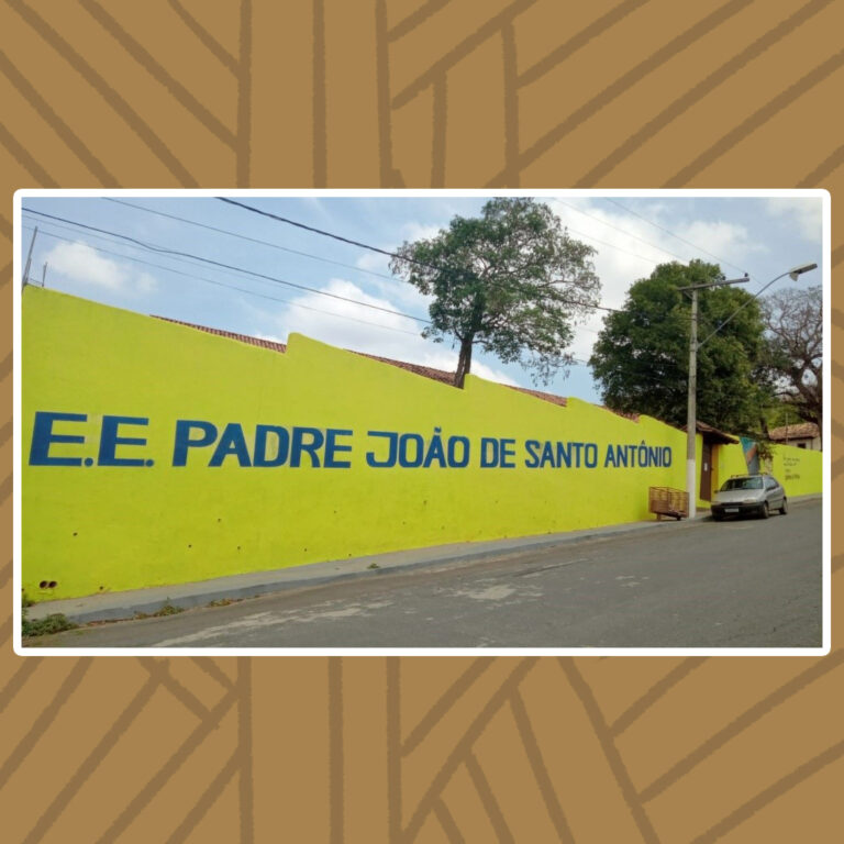 Para todos verem: Em uma rua de subida inclinada está a Escola Estadual Padre João de Santo Antônio. A escola é delimitada por um grande muro amarelo com o nome da escola escrito em letras maiúsculas azuis.