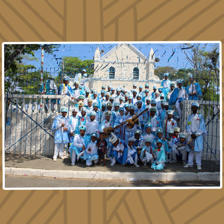 Para todos verem: Em um dia ensolarado, grupo de aproximadamente 70 membros do congado posam na escadaria da Capela de Nossa Senhora do Rosário. Os membros, em sua maioria homens, posam mais ao fundo, de pé. Alguns deles seguram instrumentos musicais. Mais à frente, crianças posam agachadas ou ajoelhadas. A vestimenta do congado é composta por manto e chapéu azul-claro, Camisa social e calça branca.