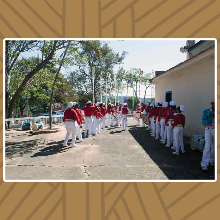 Para todos verem: Ao lado da capela, em um pátio ensolarado, cercado de árvores e mastros estendidos, o Congado do Divino se organiza em duas filas, em pares. Todos estão trajados de camisa vermelhas de mangas longas, chapéu e calças brancas, além do cinto de detalhes dourados.