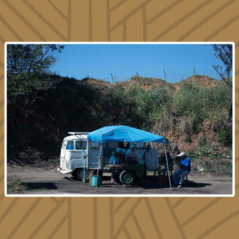 Para todos verem: Em um dia de sol forte, uma kombi branca está estacionada na beira da estrada. Suas portas laterais estão abertas, juntas a uma tenda de lona azul-clara. Dentro da tenda, uma máquina de moer cana sobre rodas. Ainda na sombra da tenda, três senhores estão sentados em banquetas de madeira.