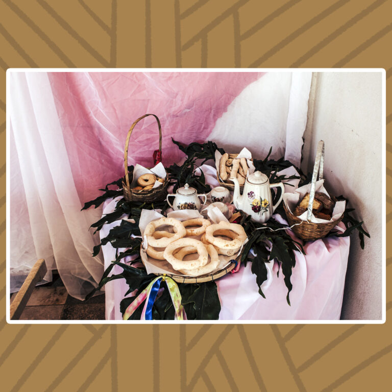 Para todos verem: Quitandas da mesa de café. A mesa é composta por cestos e peneiras de taquaras de bambu que trazem biscoitos de polvilhos redondos ou compridos, rosquinhas e bolo de fubá. Destaque ao centro para o conjunto de bule, açucareiro e xícaras de porcelana branca com detalhes florais. Flores secas pretas completam a decoração.