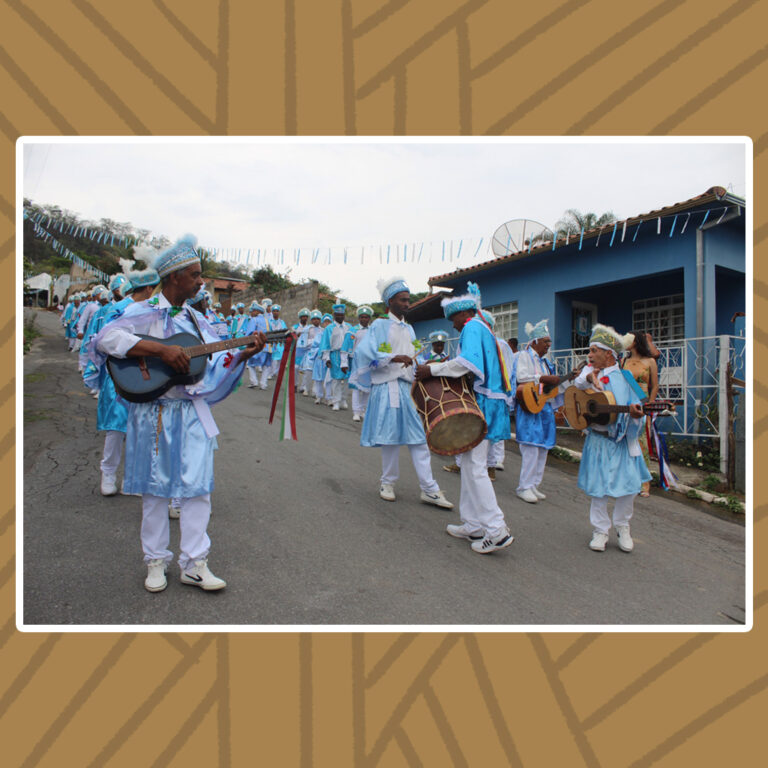Para todos verem: Em um dia nublado, o Congado de Nossa Senhora do Rosário desce uma rua íngreme. Trajados de camisa social e calças brancas, chapéus e mantos em azul-claro. Eles perfilam em pares, por duas filas. A percussão com violões e tambores puxam a procissão. A rua é decorada por um extenso varal de fitas brancas e azul-claras.