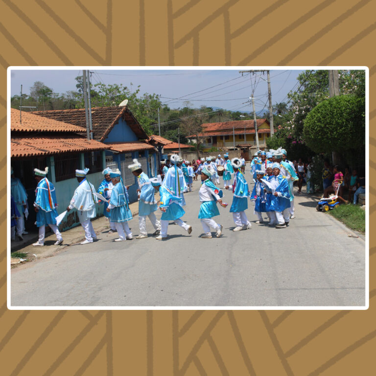 Para todos verem: Com suas vestes de camisa social e calças brancas, além do manto e chapéu azul-claros, o congado de Nossa Senhora do Rosário faz uma fila de procissão de entrada em uma casa de varanda de paredes azuis , grades e telhado inclinado. A fila faz curva no lado oposto da rua, onde o restante da população assiste ao cortejo.