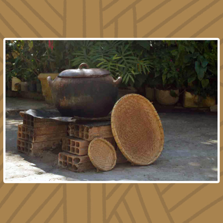 Para todos verem: Um fogão improvisado com pilhas de tijolos sustentam uma grande panela de barro. Encostada em uma das pilhas de tijolos, duas peneiras de taquara, uma pequena e a outra grande. Vasos de plantas compõem o fundo.