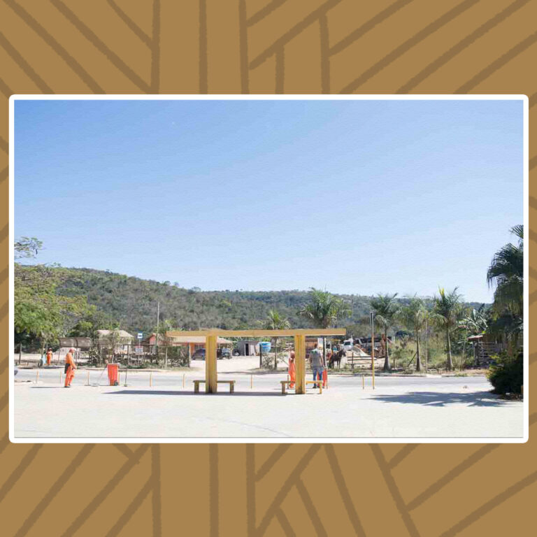 Para todos verem: Em um dia de sol forte, com vista para o morro de colinas ao fundo, está a praça principal da comunidade. A praça é uma área plana, que termina com uma margem de passeio para a estrada de asfalto. Um terminal amarelo, composto por dois pontos de ônibus em formato T, de estrutura em concreto armado, estão à margem do passeio da praça. Cada um dos pontos de ônibus possui uma pilastra de sustentação para uma laje de formato retangular, além de um banco de concreto sem encosto. Árvores variadas e palmeiras complementam a paisagem.