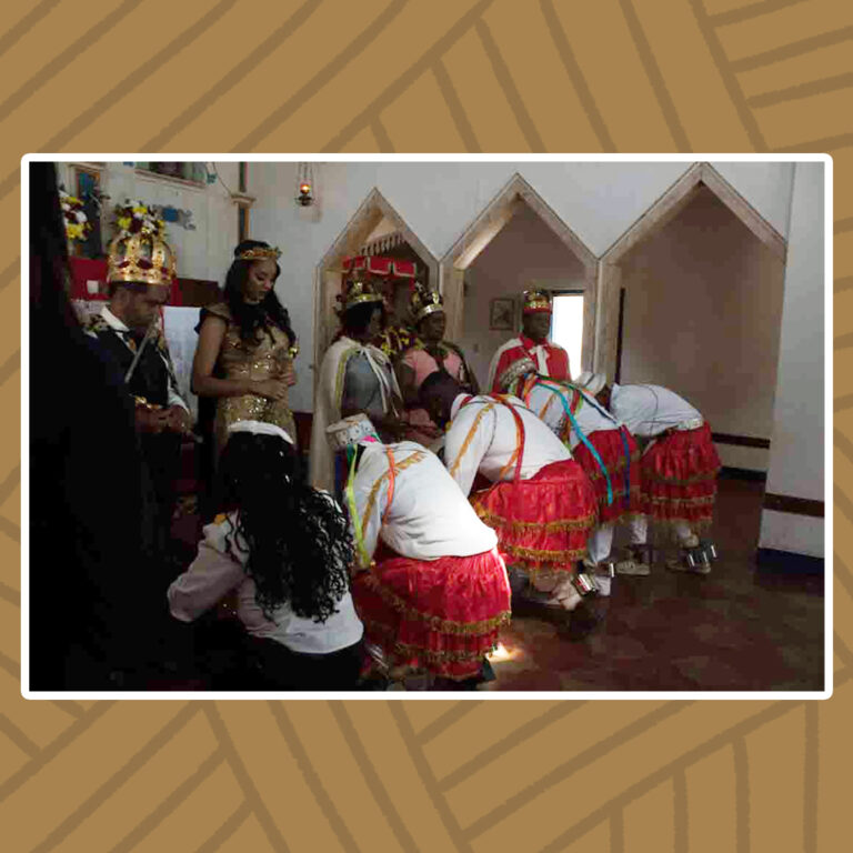 Para todos verem: Em frente ao altar mor da capela de Nossa Senhora do Rosário, o Congado realiza a cerimônia de coroação dos Reis e Rainhas do Congado. Da esquerda para a direita, sentado sobre o trono, de roupas de gala preta e grande coroa dourada sobre a cabeça, está o Rei do congado. Seguido de pé pela rainha do Congado, com suas vestes, tiara e palmas douradas. Outras duas rainhas, com coroas douradas e um Rei com coroa dourada e vestes vermelhas completam a fila. De frente para a fila e ajoelhados, estão os membros do congado fazendo reverência aos reis e rainhas. Eles trajam quepes brancos, camisas brancas de mangas longas , colares de contas e fitas multicoloridas, além de saia rodada vermelha com detalhe de franjas douradas nas pontas.