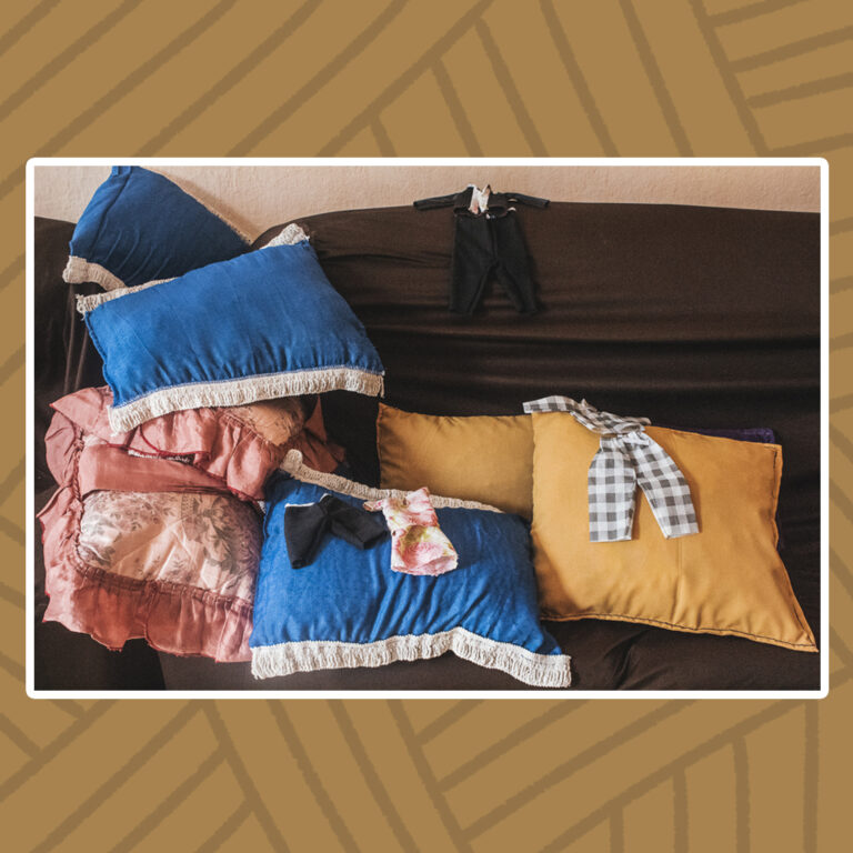 Para todos verem: Sobre sofá de tecido marrom estão estendidas peças de artesanato. Destaque para as almofadas, umas com babados brancos ou rosa, outras mais simples, nas cores azul-claro, azul-escuro, rosa e mostarda. Algumas roupas de recém-nascido, algumas feitas em tecido quadriculado, em tecido floral, outras em tecido preto, estendidas sobre as almofadas.