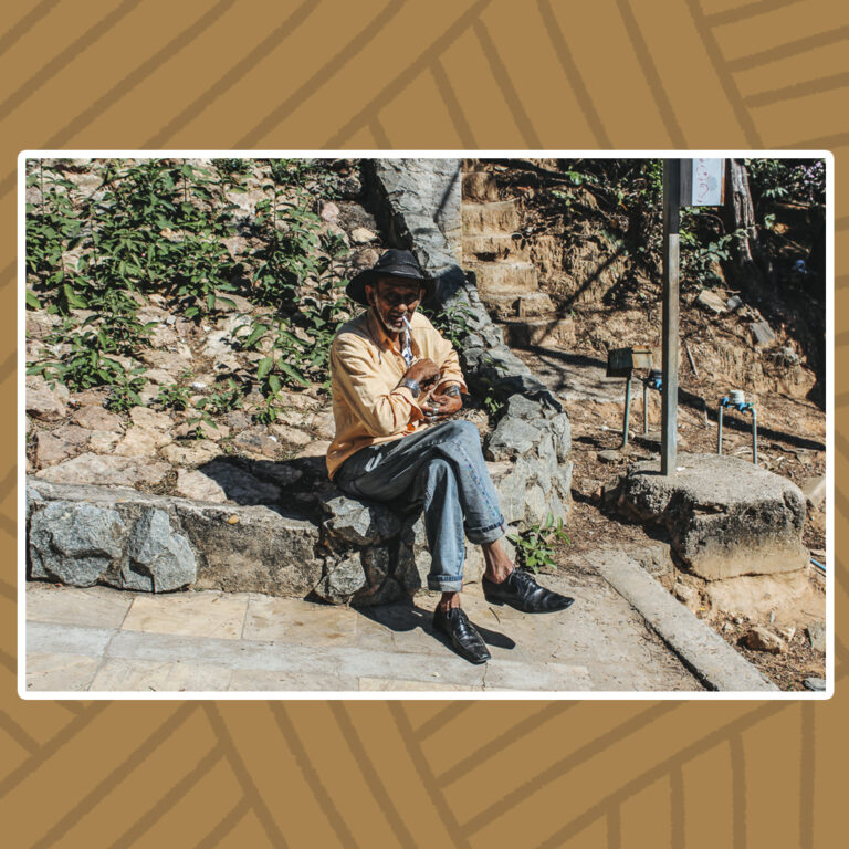 Para todos verem: Na esquina, sentado em uma mureta de pedras baixas, um senhor de pernas cruzadas fuma um cigarro. A luz do sol faz brilhar sua pele morena, contrastando com a sombra de seu chapéu preto de aba redonda. O senhor tem barba curta branca, usa camisa de manga longa em um tom laranja-claro. A calça jeans azul desbotada, com as bordas dobradas, revelam os tornozelos. Tem pulseiras nos braços e anéis prateados nos dedos. Uma das mãos procura algo no bolso de sua camisa. Seu lustroso sapato preto de bico brilha, assim como seu chapéu.
