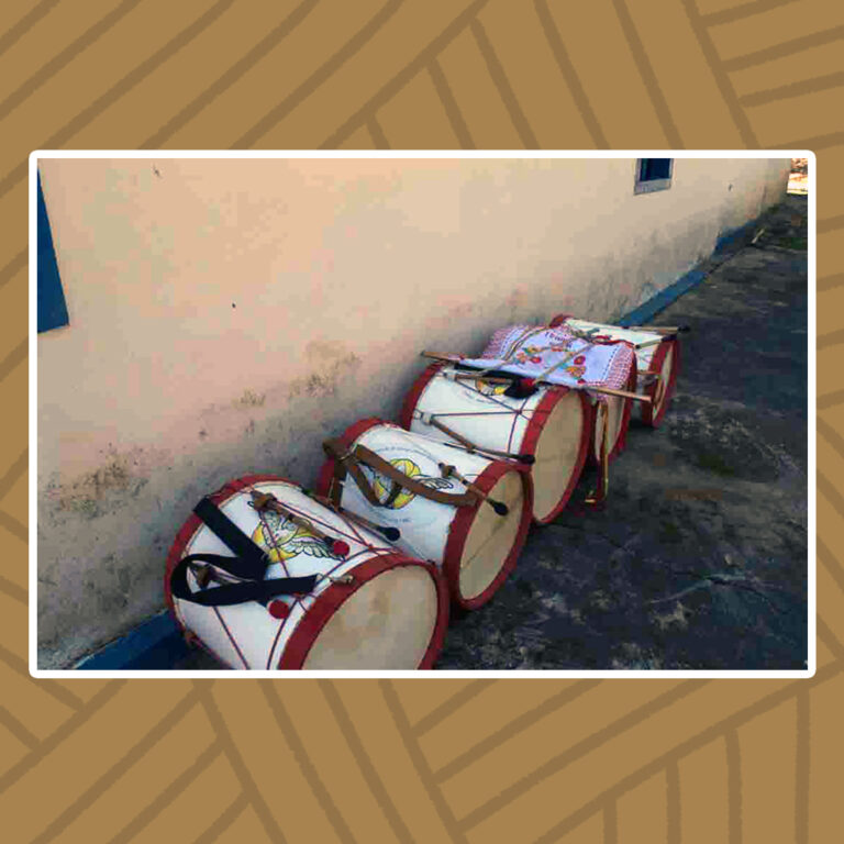 Para todos verem: Deitados, encostados em uma parede branca, cinco tambores do Congado do Divino Espírito Santo. Suas bordas são vermelhas com o topo e o fundo em branco. Seu interior também de cor branca traz a figura de uma pomba branca de asas abertas, envoltas por um esplendor de círculo amarelo.