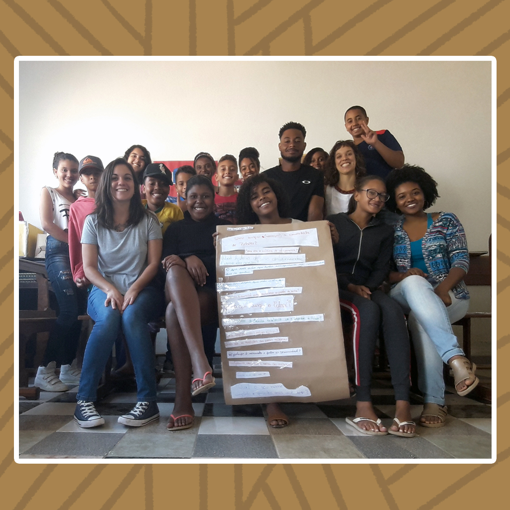 Grupo de aproximadamente 17 jovens quilombolas, alguns sentados, outros de pé, que posam para a foto. Destaque para a jovem sentada ao centro que segura um cartaz.