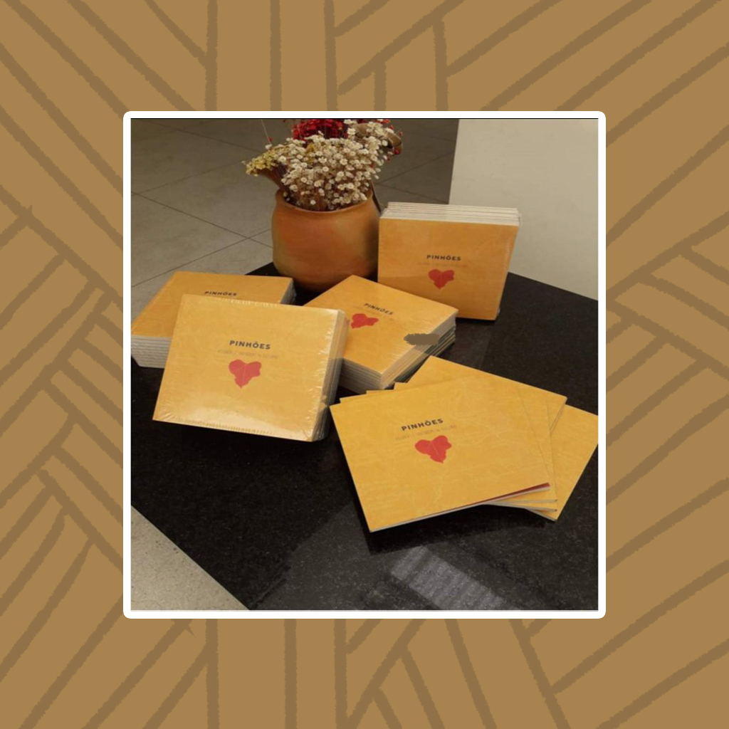 Sobre uma bancada de pedra preta estão exemplares do livro "Pinhões Histórias e Sabedorias do Quilombo". O livro possui capa amarela com título centralizado em letras maiúsculas pretas e abaixo do título, a ilustração de um pinhão vermelho. Ao fundo, um jarro com flores de capim-dourado.