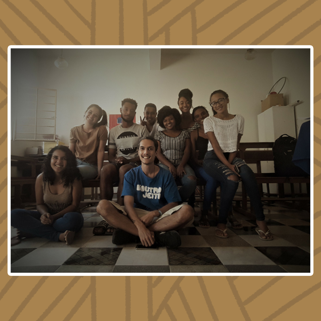 Sobre um longo banco de madeira, de um salão de paredes brancas e piso quadriculado em tons de cinza, nove jovens quilombolas posam sorridentes para a foto. Destaque para dois jovens sentados no chão, com as pernas cruzadas sendo eles uma moça de cabelos cacheados na altura dos ombros e um rapaz de cabelos cacheados, longos e presos com um nó.